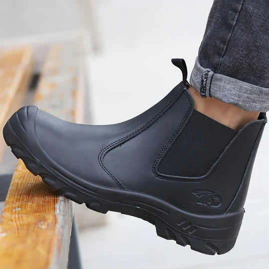 Schwarzer Chelsea-Sicherheitsstiefel aus Leder, getragen zu Jeans, Fuß auf einer Holzstufe abgestellt, Seitenansicht 1
