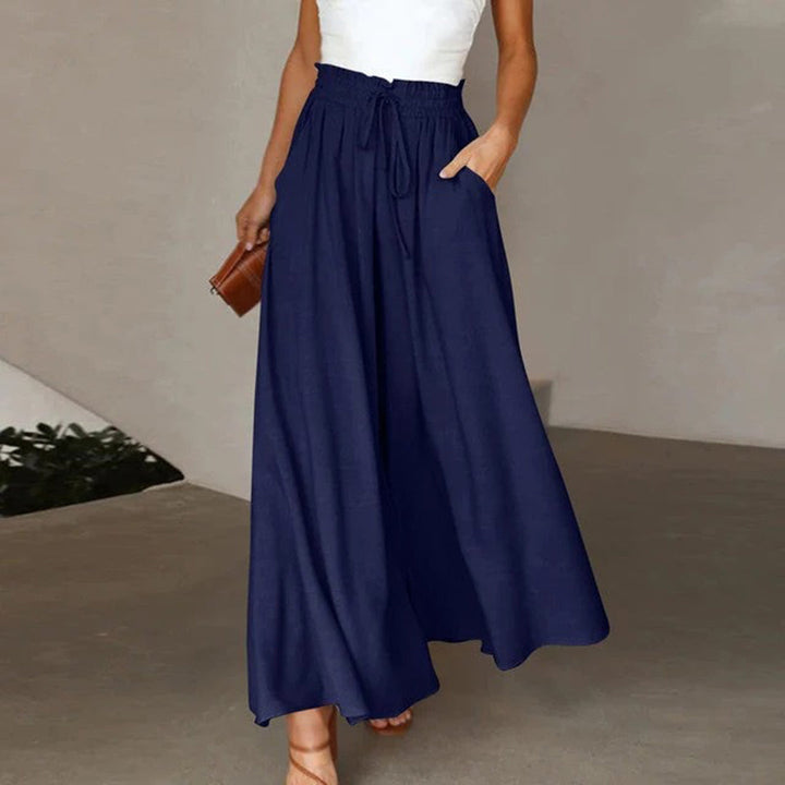 Frau in blauer, weiter Hose mit hoher Taille und weißem Top, hält braune Tasche.