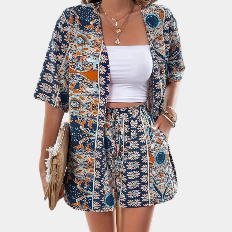Frau in blau-orange gemustertem Sommeranzug, weißem Top, mit Strohtasche und Schmuck.