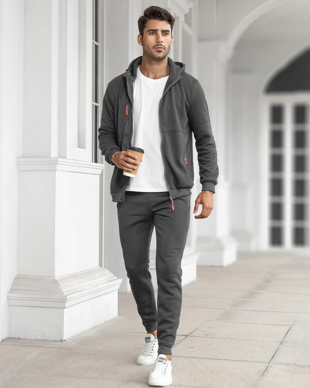 Mann im grauen Trainingsanzug mit offener Jacke über weißem Shirt, mit Kaffeebecher in einer hellen Säulenhalle 12