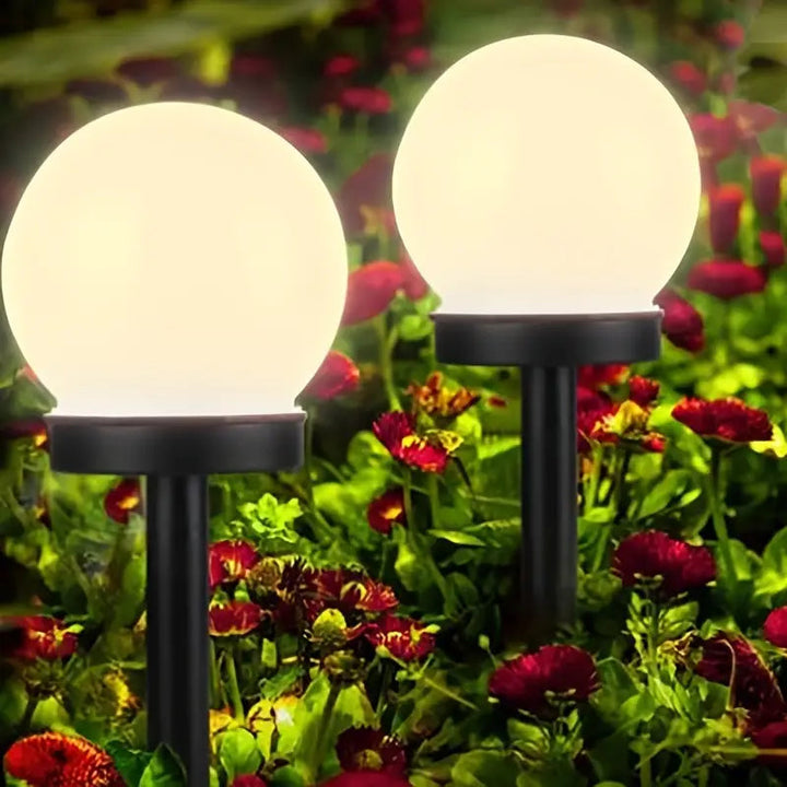 Zwei kugelförmige Solarleuchten im Garten, umgeben von roten Blumen, leuchten nachts.
