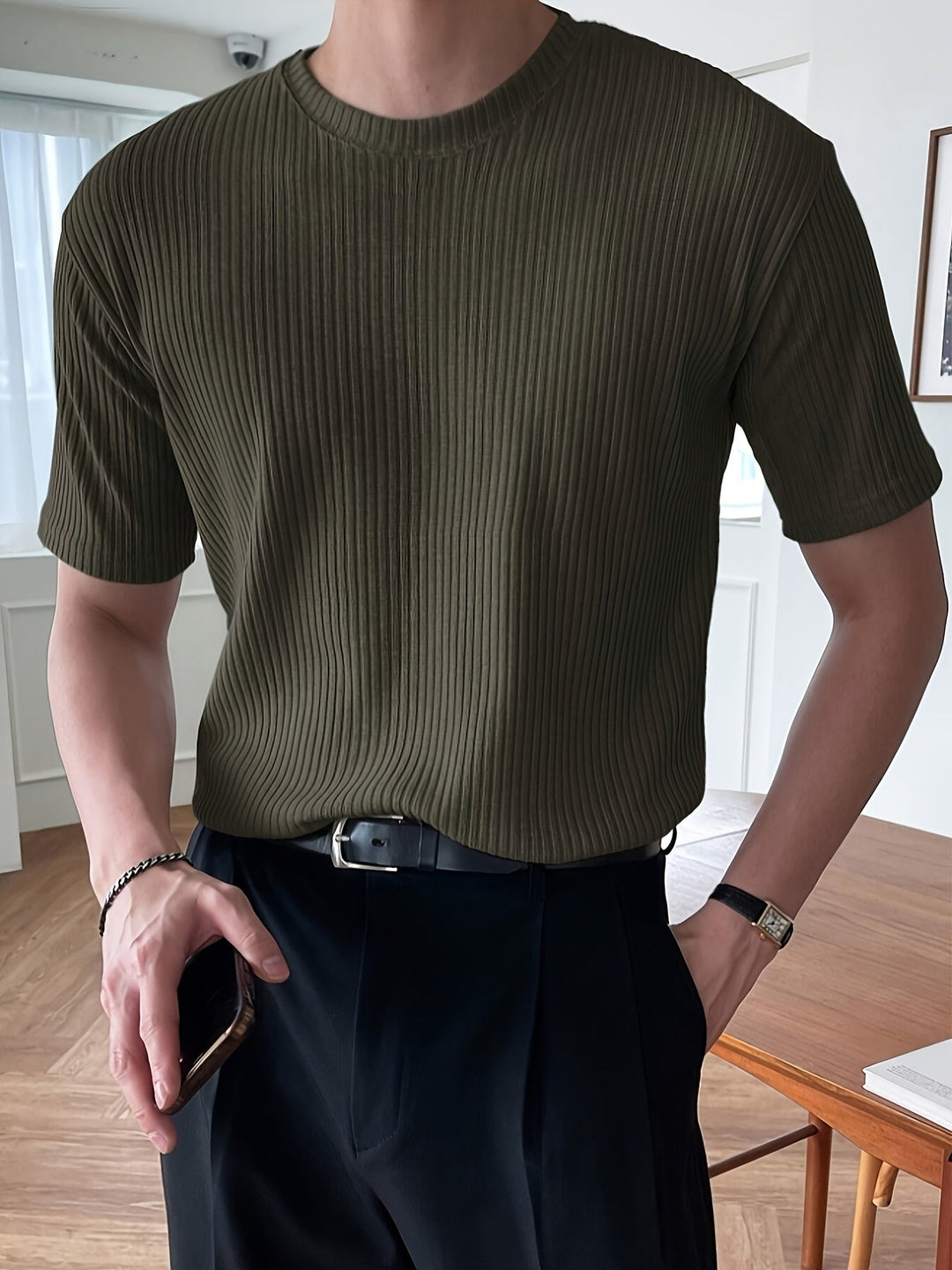Mann in olivgrünem, geripptem T-Shirt, schwarze Hose, Armbanduhr, Smartphone in der Hand.