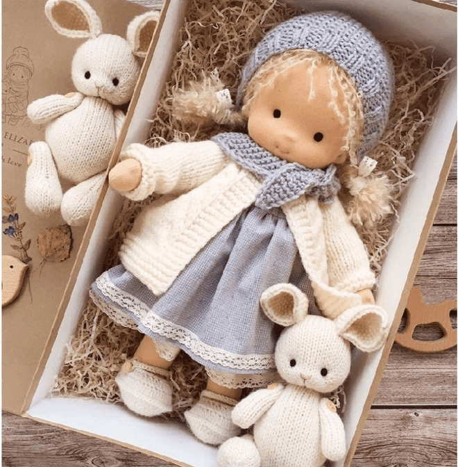 Puppe mit blauer Strickmütze, cremefarbener Jacke und Spitzenkleid im Karton, umgeben von zwei Strickhasen 3