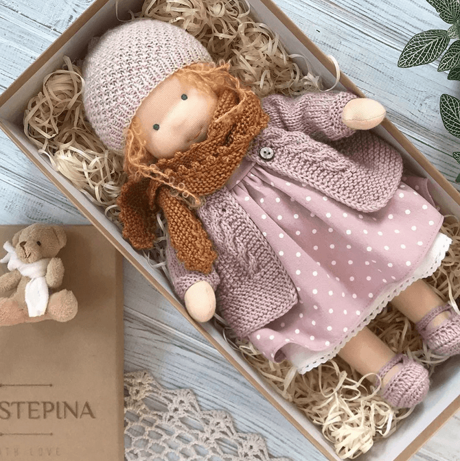Puppe mit rosa Mütze, Strickjacke und gepunktetem Kleid auf Holzwolle im Karton, daneben ein Teddy 4