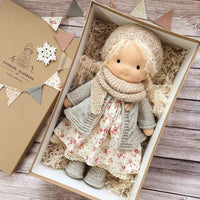 Puppe mit heller Mütze, grauer Strickjacke, Schal und Blumenkleid im geöffneten Karton mit Wimpelgirlande 6