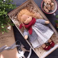 Puppe mit lockigem Haar, rotem Schal und grauer Strickjacke, im Karton liegend, daneben ein Teddy in einer Tasse 8
