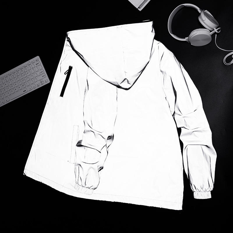 Flatlay der reflektierenden Jacke mit Kapuze und Reißverschlusstasche auf dunklem Untergrund neben Tastatur und Kopfhörern 2