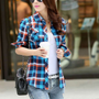 Frau in blau-kariertem Hemd, Jeans, Sonnenbrille, mit Kaffeebecher und schwarzer Handtasche.