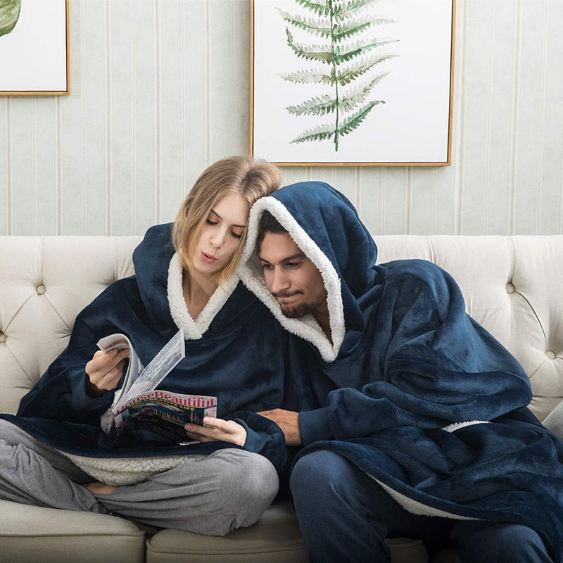 Zwei Personen in blauen Kuscheldecken mit Kapuze auf Sofa, lesen Zeitschrift, gemütlich.