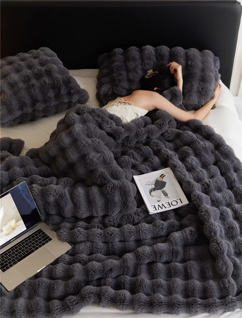 Frau liegt unter dunkelgrauer Plüschdecke mit Noppenstruktur im Bett, daneben Laptop und Magazin 2