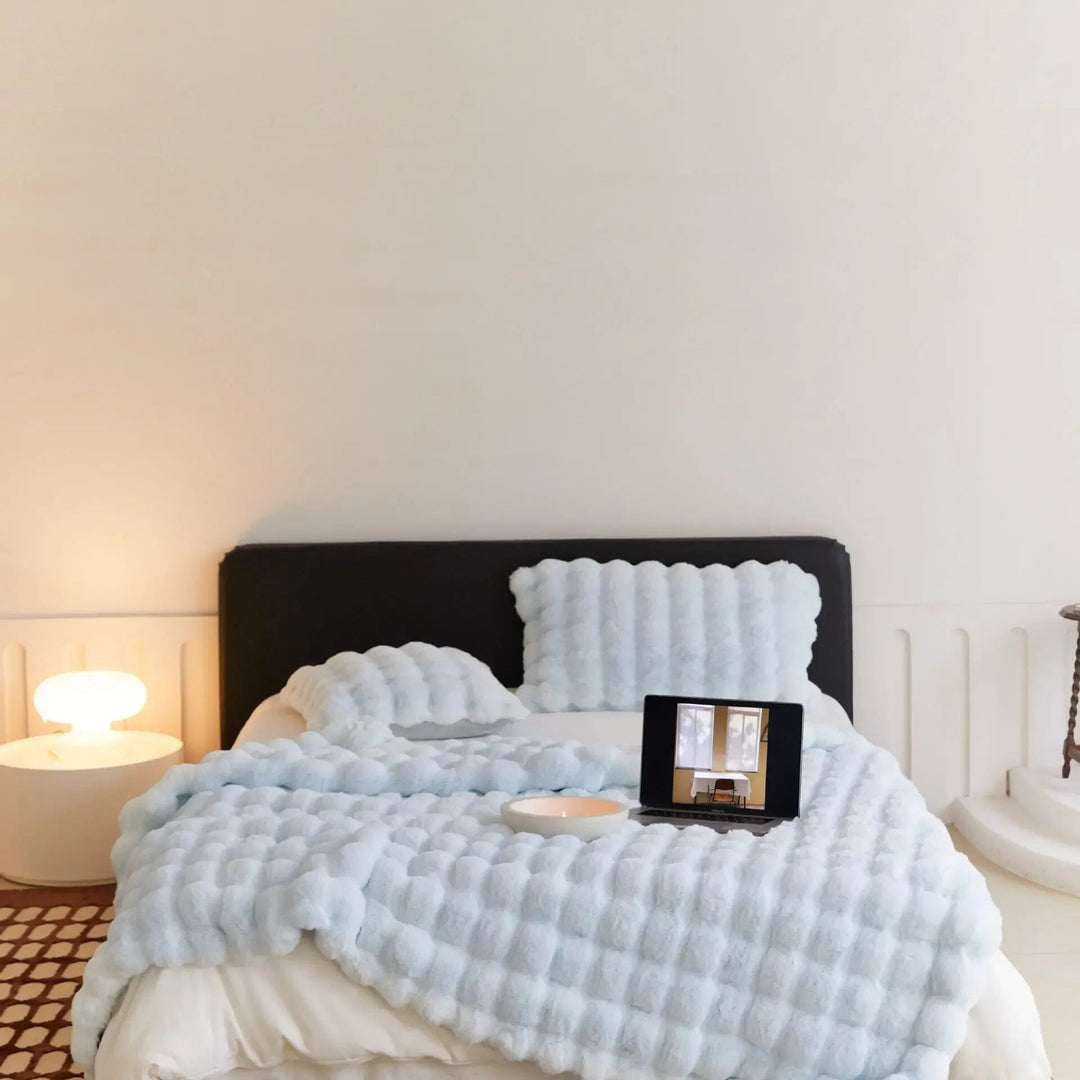 Bett mit hellblauer Noppen-Plüschdecke und passenden Kissen, darauf Laptop und Kerzenschale, Lampe links 5