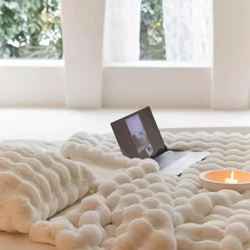 Cremeweiße Plüschdecke mit Wellenstruktur im Vordergrund, dahinter Laptop und Kerzenschale vor einem Fenster 7